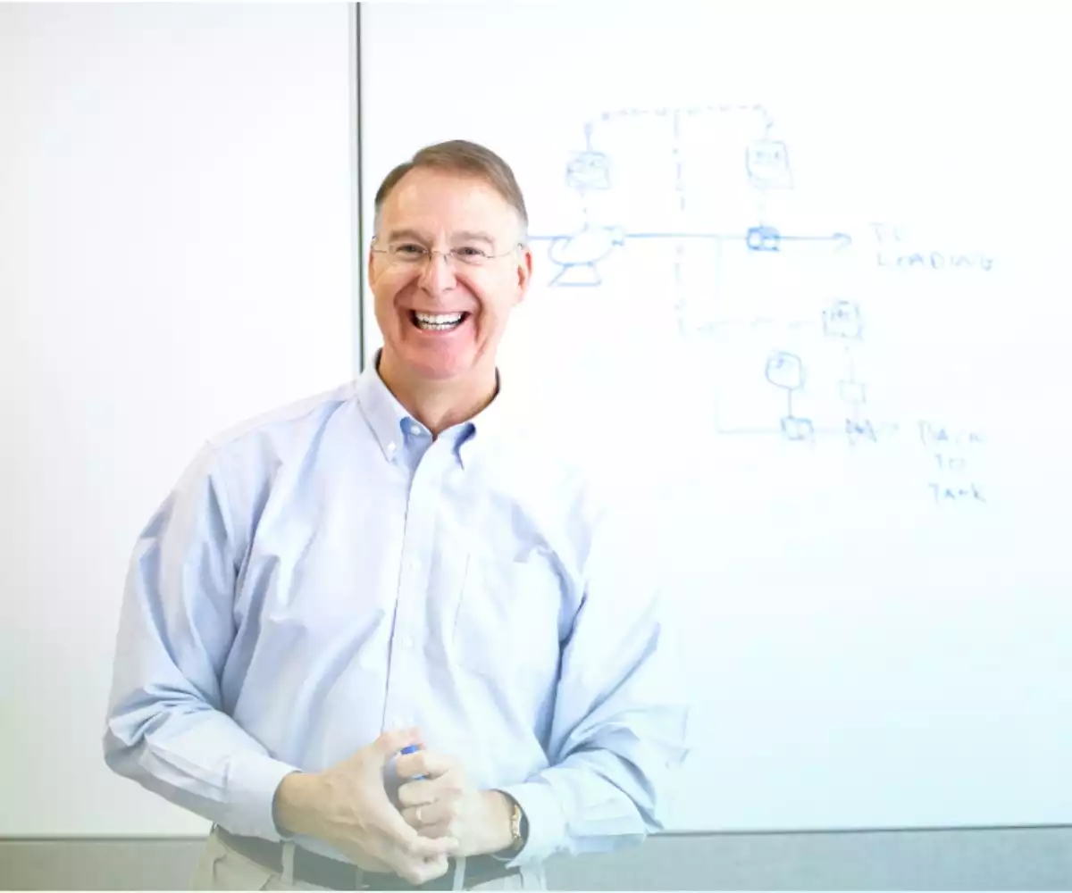 A man smiles while standing in front of a white board with diagrams.